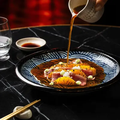 A moody and elegant shot for Akina Mumbai, featuring a tuna dish with a sauce pour. The dark marble tabletop and dramatic lighting highlight the vibrant colors of the food and the precise plating.
