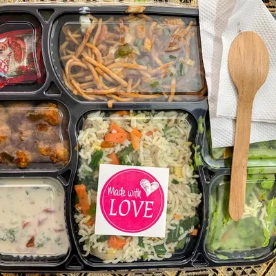 A complete meal in a box, sealed with love. This thali features my signature Veggie Pulao, Babycorn Manchurian, Hakka Noodles, Green Sauce Pasta, and a fresh salad, complete with a wooden spoon for easy eating.