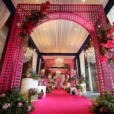 A grand entrance for a Haldi ceremony, designed with a stunning pink lattice archway and a carpeted walkway. The entire space is bathed in shades of pink and red florals, complemented by crystal chandeliers for a touch of luxe.