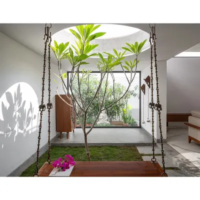 A view from the indoor swing, looking past the central courtyard tree towards a tranquil garden space with a Buddha statue. This composition highlights the layered integration of green spaces throughout the home's public areas.