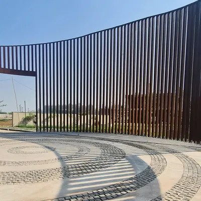 The entrance to a residential development in Raipur, featuring a gracefully curving Corten steel gate. The shadows cast by the vertical slats create a dynamic pattern on the cobblestone driveway.