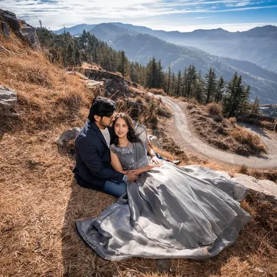 A stunning wide-angle shot of the couple nestled on a mountainside, with layers of Himalayan ranges stretching into the distance. The flowing grey gown adds to the epic feel.