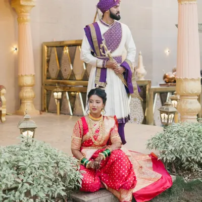 A regal portrait set against a palatial backdrop in Thane. The bride is seated in her vibrant red Nauvari saree, while the groom stands tall in his traditional kurta and pheta, creating a scene of timeless grace.