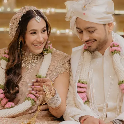 A candid, happy moment from a wedding ceremony. The bride's pastel pink lehenga is paired with a soft, doll-like makeup look, featuring rosy cheeks and a gentle glow that reflects her joy.