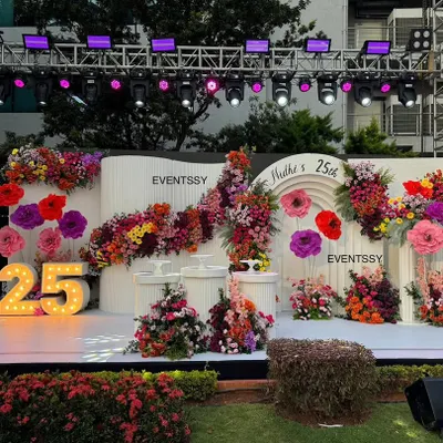 A wide shot of the stunning floral stage for a 25th birthday, featuring a clean white backdrop overflowing with colorful flowers and a large marquee number.