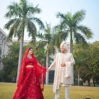 A classic wedding day portrait of the couple holding hands in a beautiful garden. The bride's vibrant red lehenga and the groom's elegant white sherwani create a timeless image.