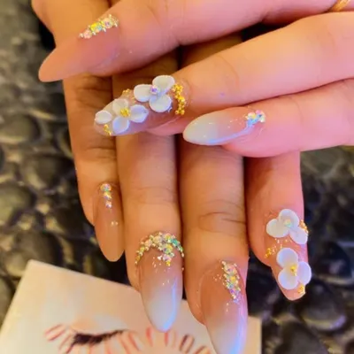 An intricate design featuring ombre nail extensions with sculpted 3D white flowers. Tiny crystals and glitter beads are added at the base and tips for a delicate, sparkling finish.