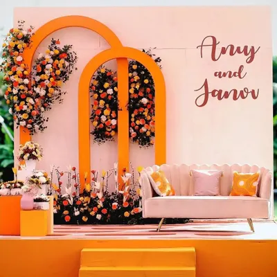 A vibrant and modern stage setup for an engagement party. The bold orange arches, combined with cascading marigolds and a chic scalloped sofa, create a fresh and joyful look.