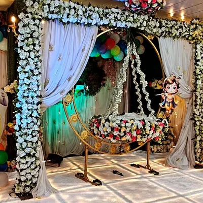 A gorgeous Krishna-themed naming ceremony decoration. The setup includes a decorated cradle under a floral arch, character cutouts, and an LED-lit stage, creating a blessed and beautiful environment for the 'palna' ceremony.
