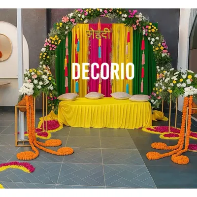 This colorful mehndi seating area features a backdrop of bright pink and green drapes against a floral arch. The low seating with cushions provides a comfortable and traditional space for the bride during the ceremony.