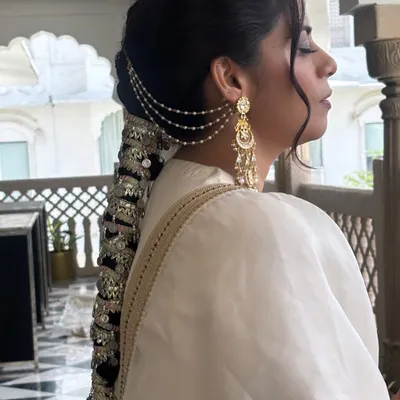 A stunning side profile of an intricate bridal braid. This style features a traditional Punjabi parandi with mirror work and ghungroos, complemented by delicate pearl chains that connect to the earrings. It's a true work of art.