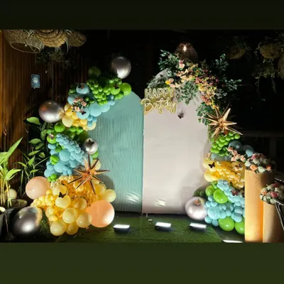 This elegant birthday setup features a unique house-shaped backdrop with floral arrangements. The organic balloon garland in shades of green, yellow, and peach adds a soft and sophisticated touch to the decor.