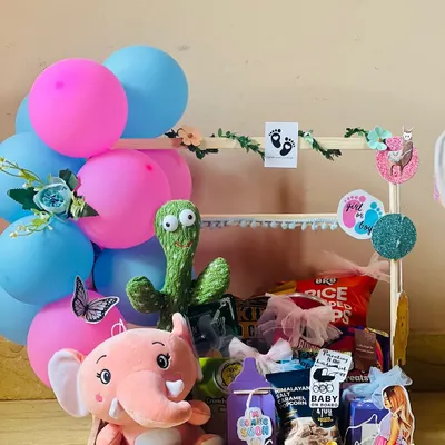 This baby shower hamper is full of fun and surprises. It features a pink elephant and a dancing cactus toy, along with snacks and treats, all set in a decorated wooden cart perfect for a party centerpiece.