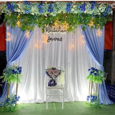 An elegant stage decoration for a 'Khitanan' ceremony, featuring blue and white drapes, floral arrangements, and fairy lights.