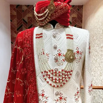 A classic white sherwani designed for a royal wedding look. It features red floral embroidery, a matching red turban, and a multi-layered pearl and ruby necklace, all put together for a complete groom's outfit.