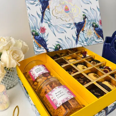 Our signature Diwali hamper in a vibrant yellow and blue box, featuring a beautiful parrot design. Inside, you'll find jars of savory crackers and sweet medley, alongside compartments of fresh brownie bites and other baked goods.