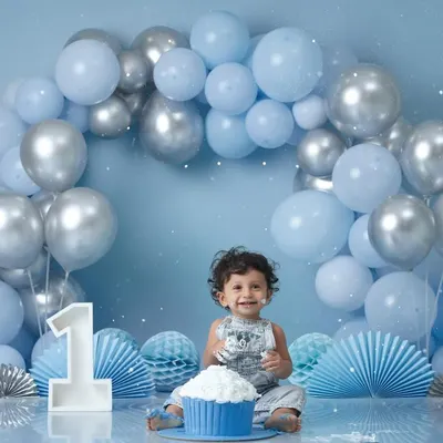 A wide shot of a baby boy's first birthday cake smash, showing him sitting happily in front of a large blue and silver balloon arch.