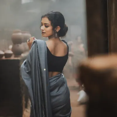 A beautiful back profile shot that creates a sense of mystery and elegance. The model, draped in a simple grey saree, looks over her shoulder in a rustic, hazy setting, highlighting her graceful posture.