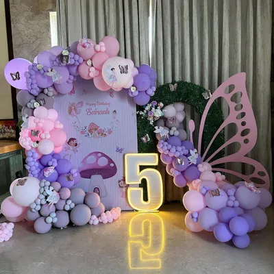 A whimsical fairy and butterfly-themed fifth birthday setup, featuring a large light-up number '5' and a stunning pink butterfly wing prop. The scene is completed with a custom backdrop of an enchanted forest and a pastel balloon arch in pink, purple, and grey.