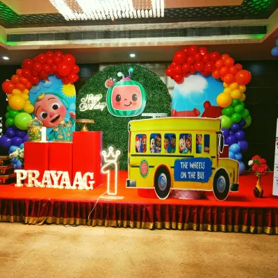 A vibrant Cocomelon themed first birthday party. The stage setup includes a large school bus prop, character cutouts, and a colorful balloon arch over a grass-textured backdrop.