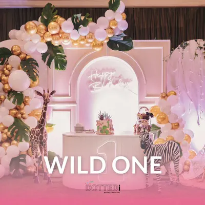 A "Wild One" jungle-themed first birthday party setup. The decor features a beautiful balloon arch in white and gold, life-sized giraffe and zebra figures, and a custom cake table.