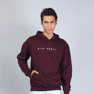 A confident, front-facing shot of a model in a maroon "Rise Above" hoodie. The clean grey background ensures the focus remains entirely on the product.