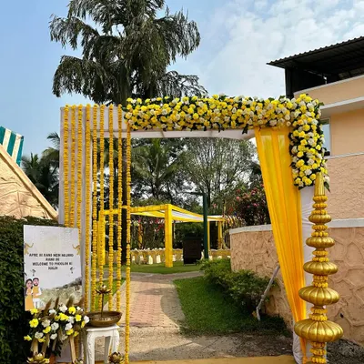 A bright and welcoming entrance for a Haldi ceremony, featuring a simple arch decorated with yellow marigold garlands and a custom welcome sign. The golden temple-style pillar adds a touch of traditional elegance to the outdoor venue.