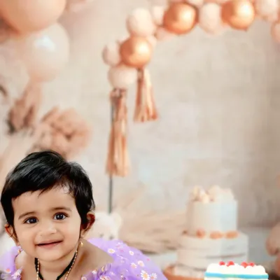 A beautiful baby girl with a radiant smile, captured during her cake smash session with a pastel balloon arch in the background.
