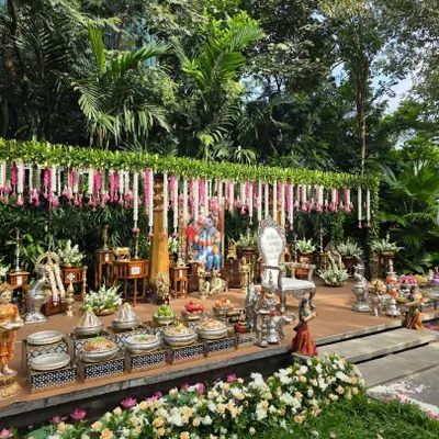 A beautiful outdoor stage set against a lush green backdrop of trees. The decor features a canopy of leaves with hanging pink and white garlands, and a Ganesha idol at the center.
