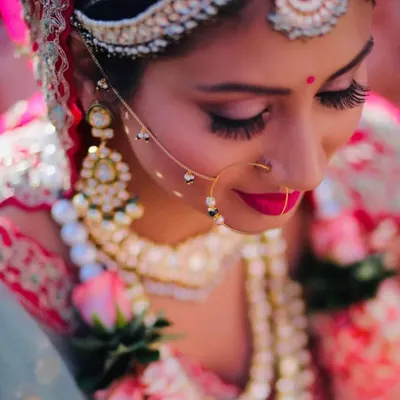 A detailed close-up of a bride's makeup, featuring dramatic winged eyeliner, long lashes, and a bold pink lip. The look is perfectly coordinated with her traditional matha patti and nath.
