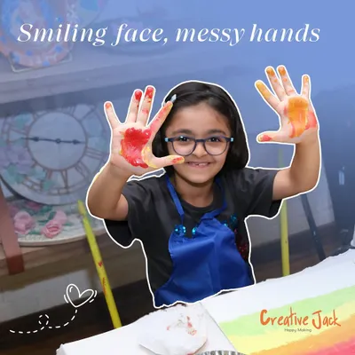 A young girl with glasses smiles, showing off her paint-covered hands. This is the sign of a fun and successful art session.