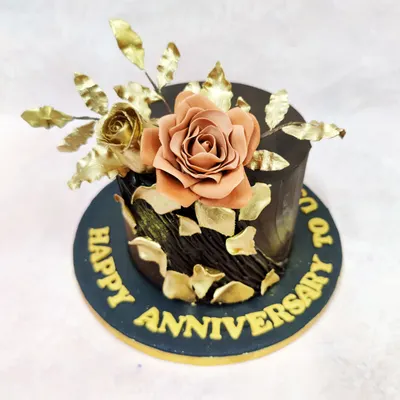 This anniversary cake is a statement of opulent romance, featuring a rich, dark chocolate exterior with a textured finish. A handcrafted sugar rose in a muted blush tone is complemented by gilded leaves and shimmering gold leaf fragments, creating a luxurious and timeless design.