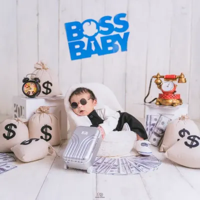 A series from a 'Boss Baby' themed newborn shoot. The setup includes a tiny armchair, a miniature briefcase, and prop money, creating a playful and humorous scene for a baby boy's first portraits.
