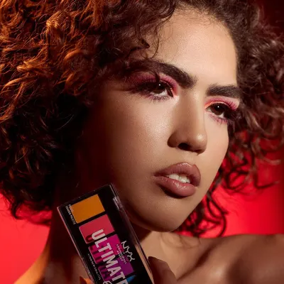 A powerful and moody shot featuring a model with vibrant pink eyeshadow, holding a NYX Ultimate Edit Palette. The dramatic red lighting and her intense expression create an edgy, editorial feel, perfect for a brand that champions bold self-expression through makeup.