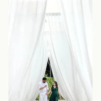 A stunning pre-wedding shot of a couple walking through a corridor of flowing white curtains. This setup creates a dreamy and elegant atmosphere, perfect for a romantic and visually striking photograph.