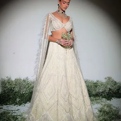 Embodying timeless elegance, this pristine ivory lehenga is embellished with delicate pearls and intricate craftsmanship. The look is completed with feather-trimmed cape sleeves for a touch of modern grace.