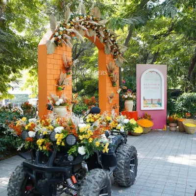 A grand entrance for an orange-themed Haldi, featuring a flower-adorned ATV for the groom's entry. The orange brick-textured arch is decorated with pampas grass and marigolds.