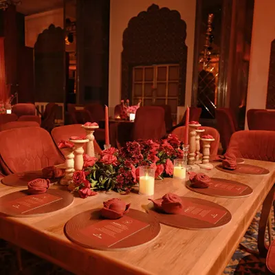A long wooden table set for an intimate dinner, surrounded by plush red velvet chairs. The centerpiece of red roses and candles creates a warm and romantic focal point.