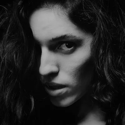An intense black and white close-up. The single light source creates a dramatic shadow across the face, highlighting the eye and creating a sense of mystery and power.