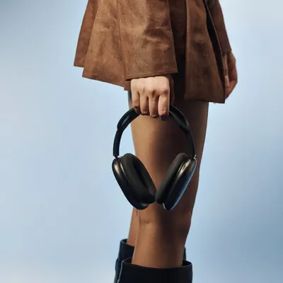 A clean, modern product-in-context shot. The focus is on the sleek design of the black headphones, held against a simple brown skirt and blue background. This style is perfect for e-commerce and social media campaigns for modern accessories.