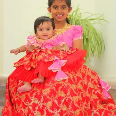 Twinning sisters, one big and one small, in our custom-made outfits. The older sister wears a pink blouse, while the baby wears a matching layered pattu pavadai in a vibrant red Banarasi silk.