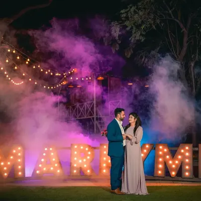 A grand romantic gesture set against our "MARRY ME" marquee letters. The combination of purple smoke, string lights, and the rustic cabin backdrop makes for an unforgettable proposal or pre-wedding photograph.