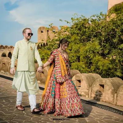 A beautiful shot of a couple in vibrant, traditional attire walking hand-in-hand along a historic fort wall. This image captures the essence of a royal Rajasthani pre-wedding shoot, filled with color and heritage.