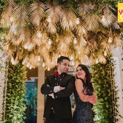 The happy couple, Rucha and Chinmay, pose under a stunning canopy of golden leaves and Edison bulbs at their tropical-themed Sangeet night.