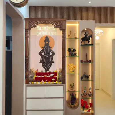 This pooja unit is designed as a statement piece, featuring a traditionally carved wooden frame around a backlit panel. It includes drawers for storage and an illuminated glass cabinet on the side for displaying religious artifacts.