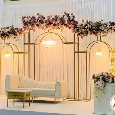 Here is a clear view of a modern and elegant stage I designed. The setup features geometric gold frames, a chic white lounge sofa, and a beautiful arch of purple and pink flowers. A delicate chandelier hangs in the center, adding a touch of classic luxury to the contemporary design.