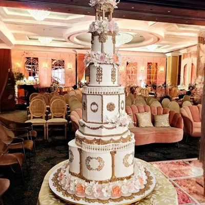 A full view of our architectural engagement cake, standing tall in a beautifully decorated ballroom. The design features multiple tiers with distinct shapes, ornate gold detailing, and a floral gazebo topper, creating an unforgettable presence.