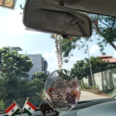This custom car hanging with Ayat ul Kursi is shown in its natural setting, hanging from a rearview mirror. It includes real flowers and glitter, catching the sunlight beautifully.