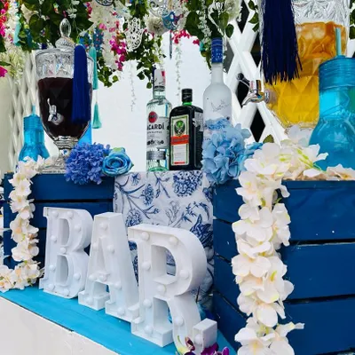 A beautiful overview of the Santorini-themed bar. The setup features a 'BAR' marquee sign, blue and white decor, floral garlands, and large glass dispensers filled with colorful cocktails, creating a stunning visual.