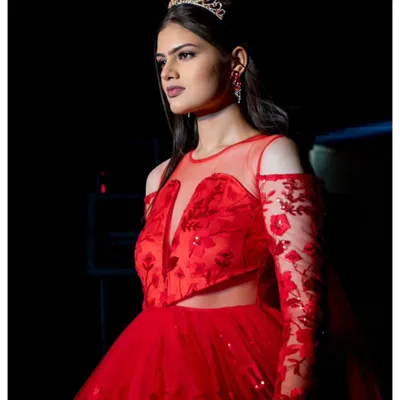 A princess-style red gown captured in its full glory on the runway. The lighting highlights the sheer, embroidered sleeves and the sparkle of the tiara, creating a fairy-tale moment during the fashion show.